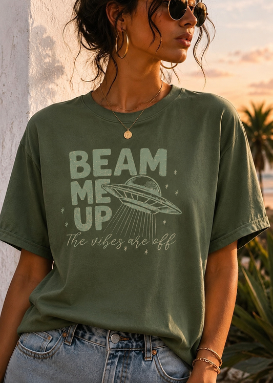 A woman wearing a faded olive green graphic t-shirt with a light green print that reads 'BEAM ME UP The vibes are off' alongside a UFO illustration. She is wearing blue jeans and sunglasses, standing outdoors against a textured white wall during sunset.