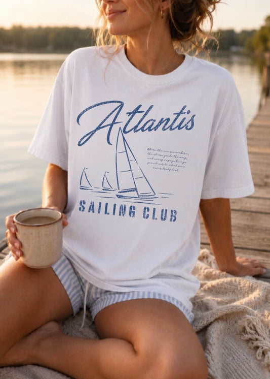 A woman sitting on a dock by the water wearing a white graphic T-shirt with a blue sailboat design and the words 'Atlantis SAILING CLUB'. She is holding a mug and wearing striped shorts.