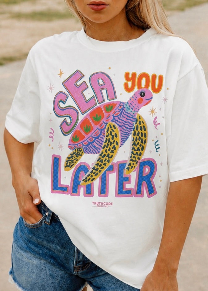 Person wearing a white t-shirt with a colorful turtle graphic and text on a beach. Text says Sea You Later.