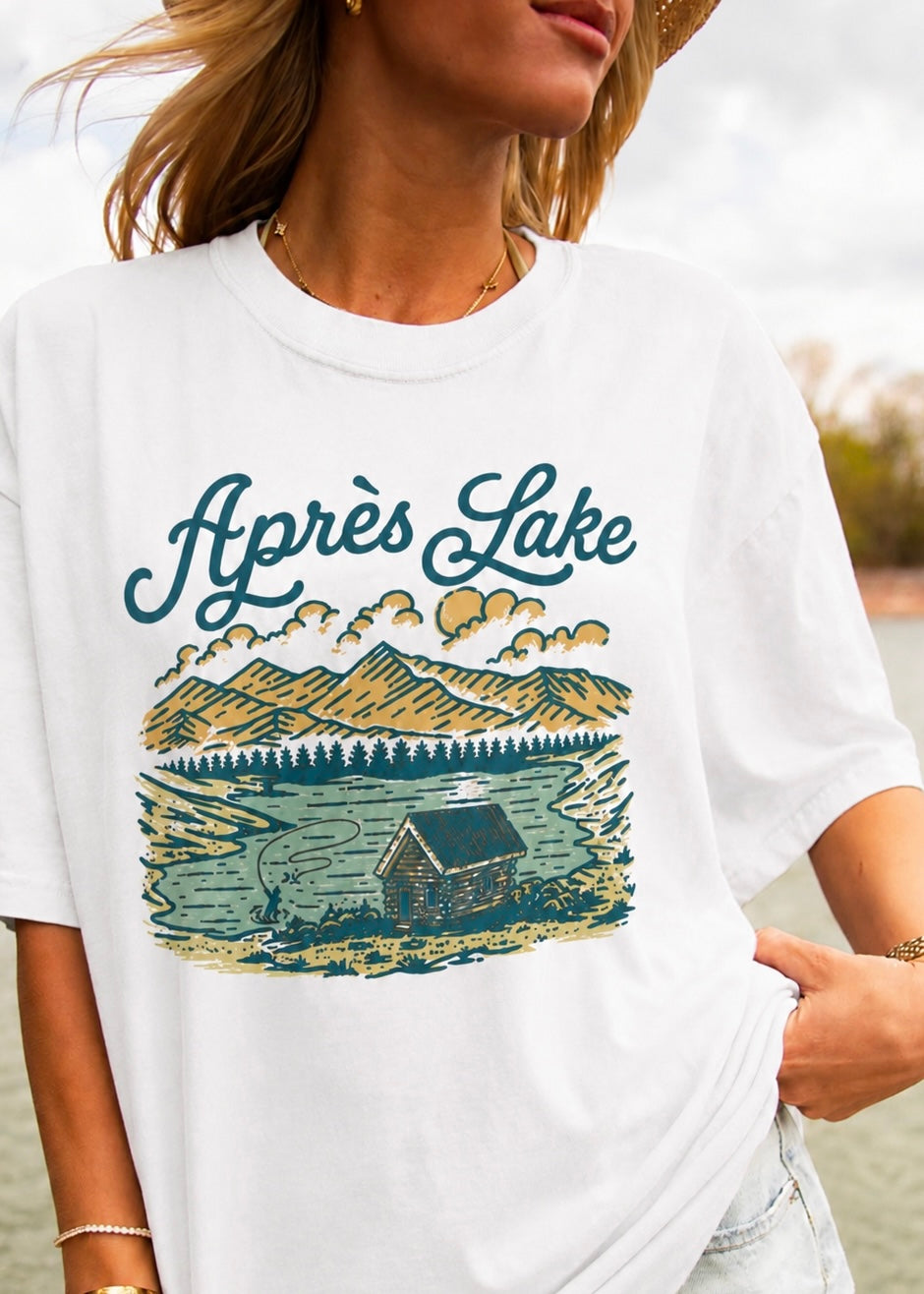 A woman wearing a white graphic t-shirt featuring a vintage-style illustration of a cabin by a lake with the text 'Après Lake' in teal script.