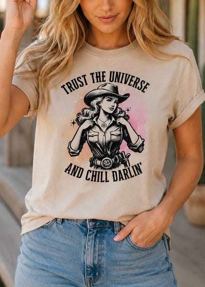 A woman wearing a beige graphic t-shirt with a western-themed print of a cowgirl and the text 'TRUST THE UNIVERSE AND CHILL DARLIN'', paired with blue jeans.