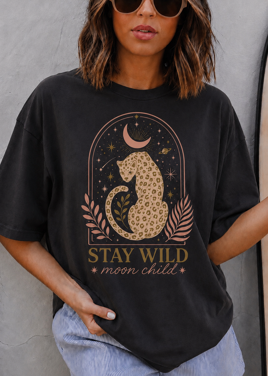 A woman wearing a black graphic t-shirt with a cat design and the text 'STAY WILD moon child', paired with light blue jeans, set against a gray concrete wall.