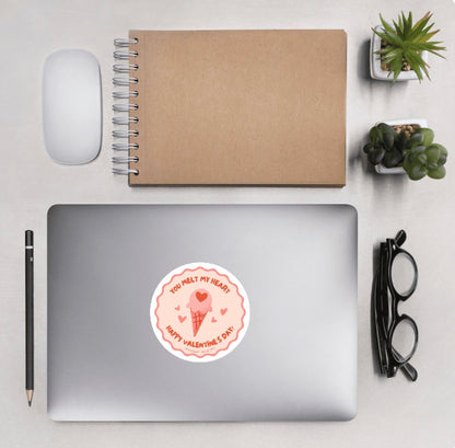 Laptop with a sticker of an ice cream cone graphic design and text 'You melt my heart, Happy Valentine's Day', background of aerial view of desk and accesories.