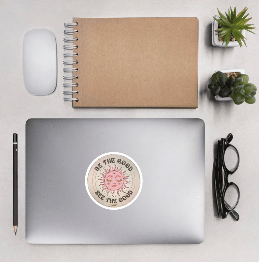 Laptop showing a sticker with text 'Be the Good, See the Good', on a desk with desktop accessories
