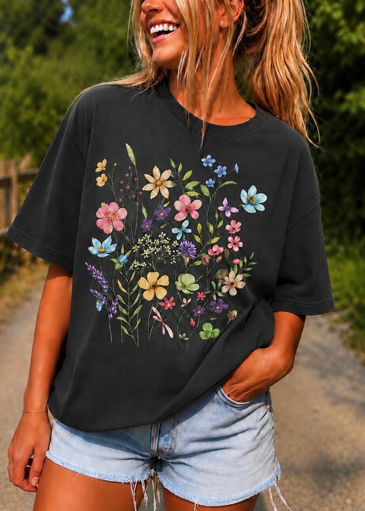 A woman wearing a black short-sleeve t-shirt with a colorful floral print on the front, paired with light blue denim shorts, standing outdoors on a path with green foliage in the background.