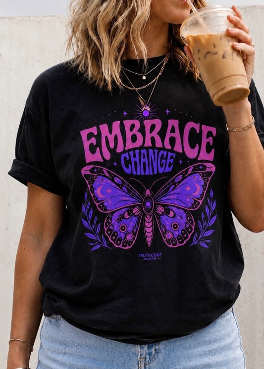 Person wearing a black t-shirt with a bright and colorful purple and pink butterfly graphic and 'Embrace Change' text, holding a drink.