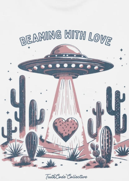 Close view of white sweatshirt with a heart in a UFO beam and desert scape and text 'Beaming with Love' 
