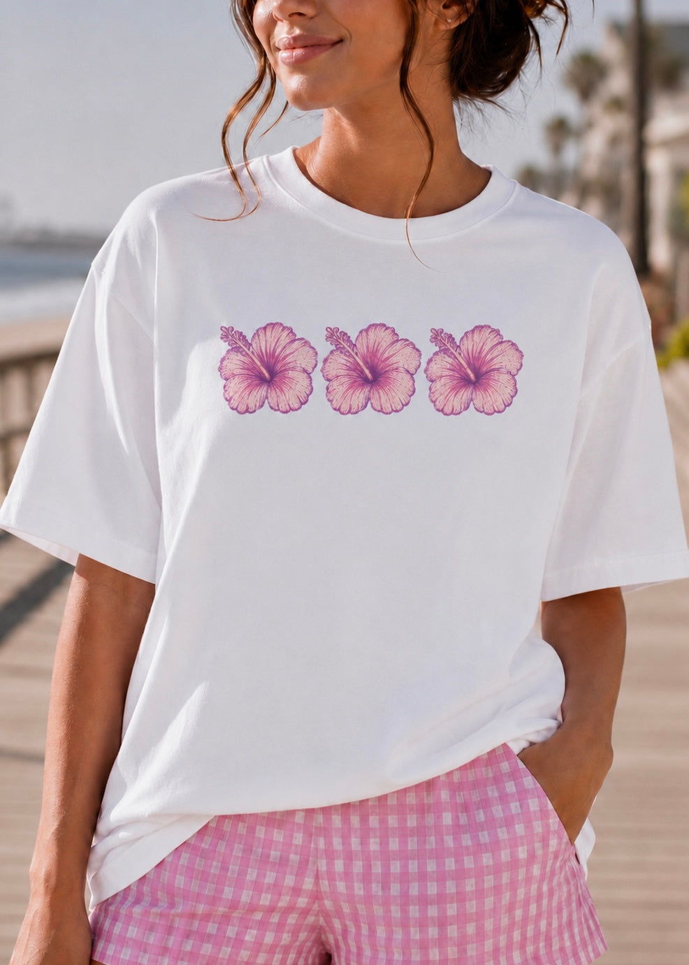 A woman wearing a white short-sleeve crewneck t-shirt with a graphic of three pink hibiscus flowers across the chest, paired with pink and white gingham shorts, set against a blurred outdoor background.