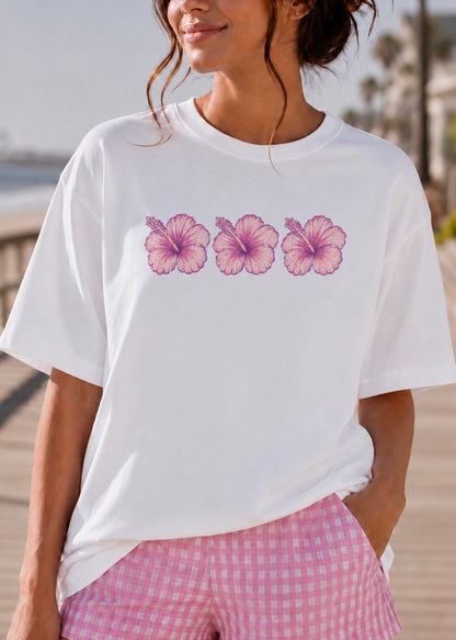 A woman wearing a white short-sleeve crewneck t-shirt with a graphic of three pink hibiscus flowers across the chest, paired with pink and white gingham shorts, set against a blurred outdoor background.