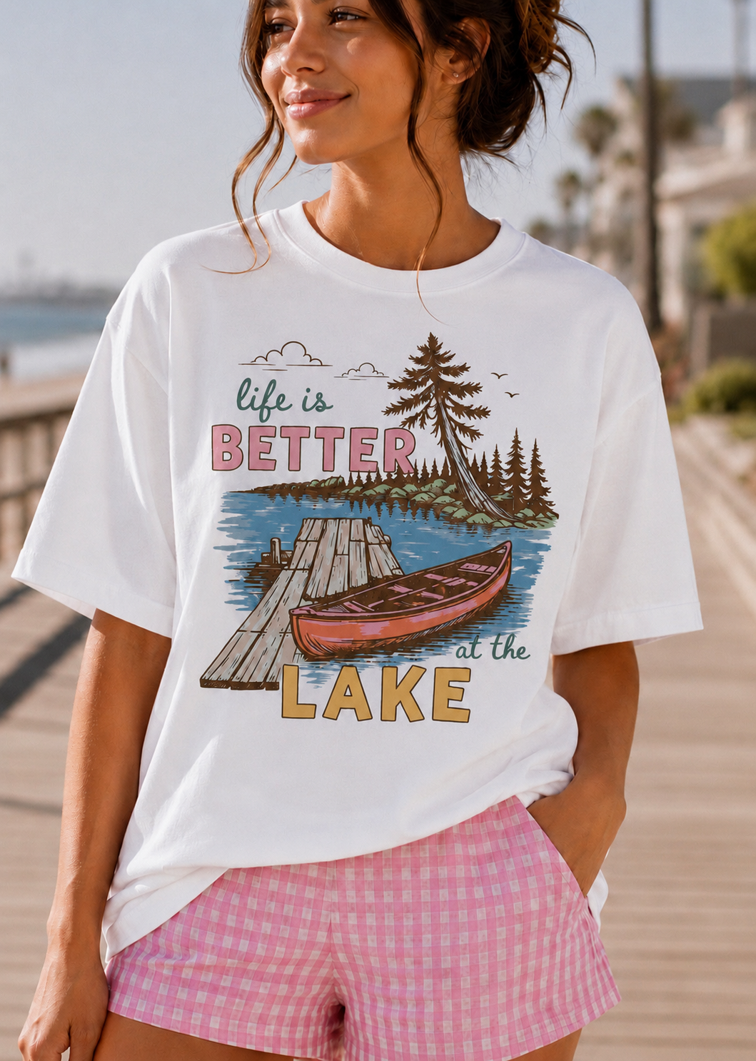 A woman wearing a white graphic t-shirt with a 'life is BETTER at the LAKE' design featuring a wooden dock, pine trees, and a red canoe, paired with pink gingham shorts, set against a sunny boardwalk background.