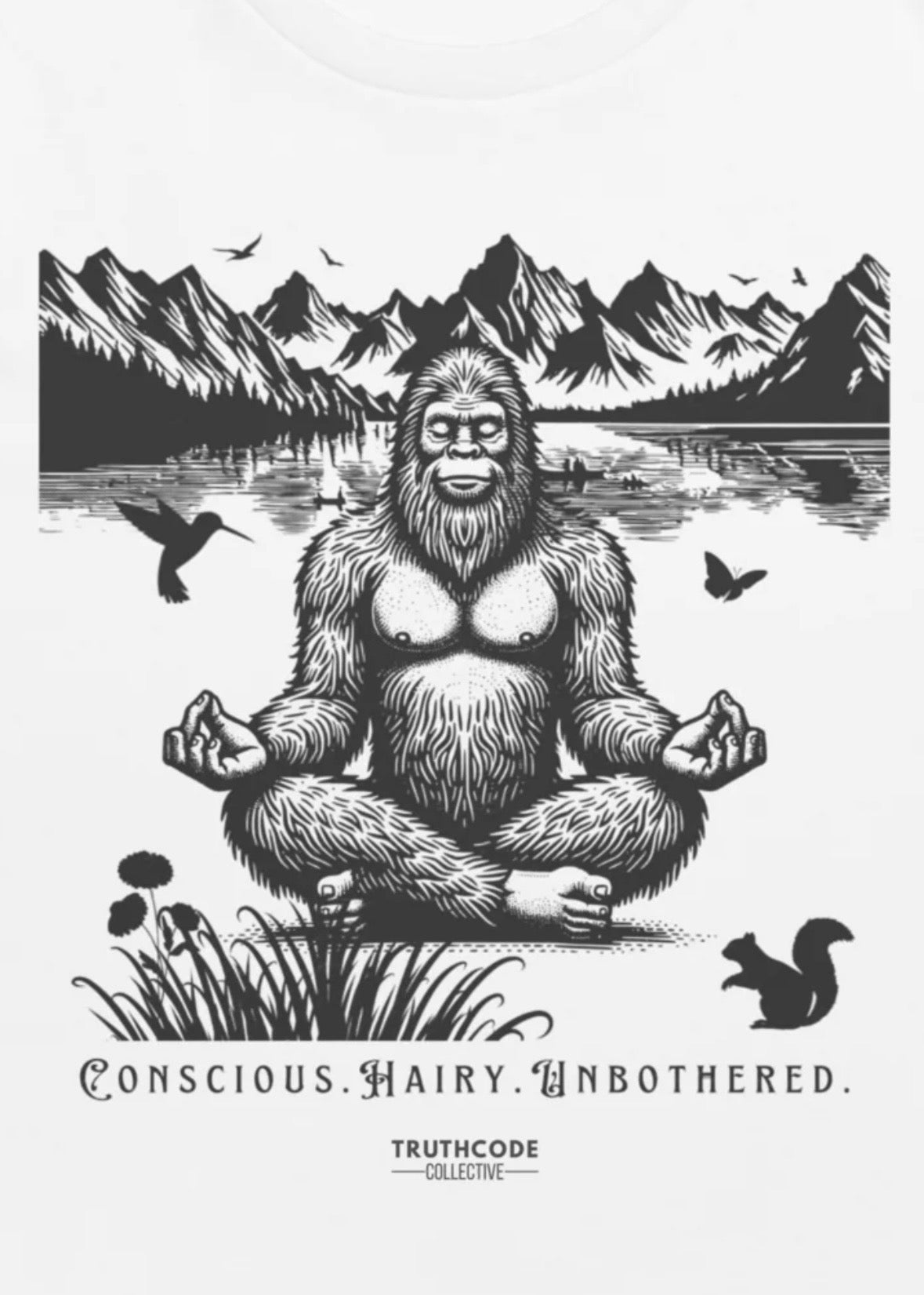 Close view of a white t-shirt with a graphic of a meditating Sasquatch by a lake with mountains in the background, featuring the text 'Conscious. Hairy. Unbothered.'