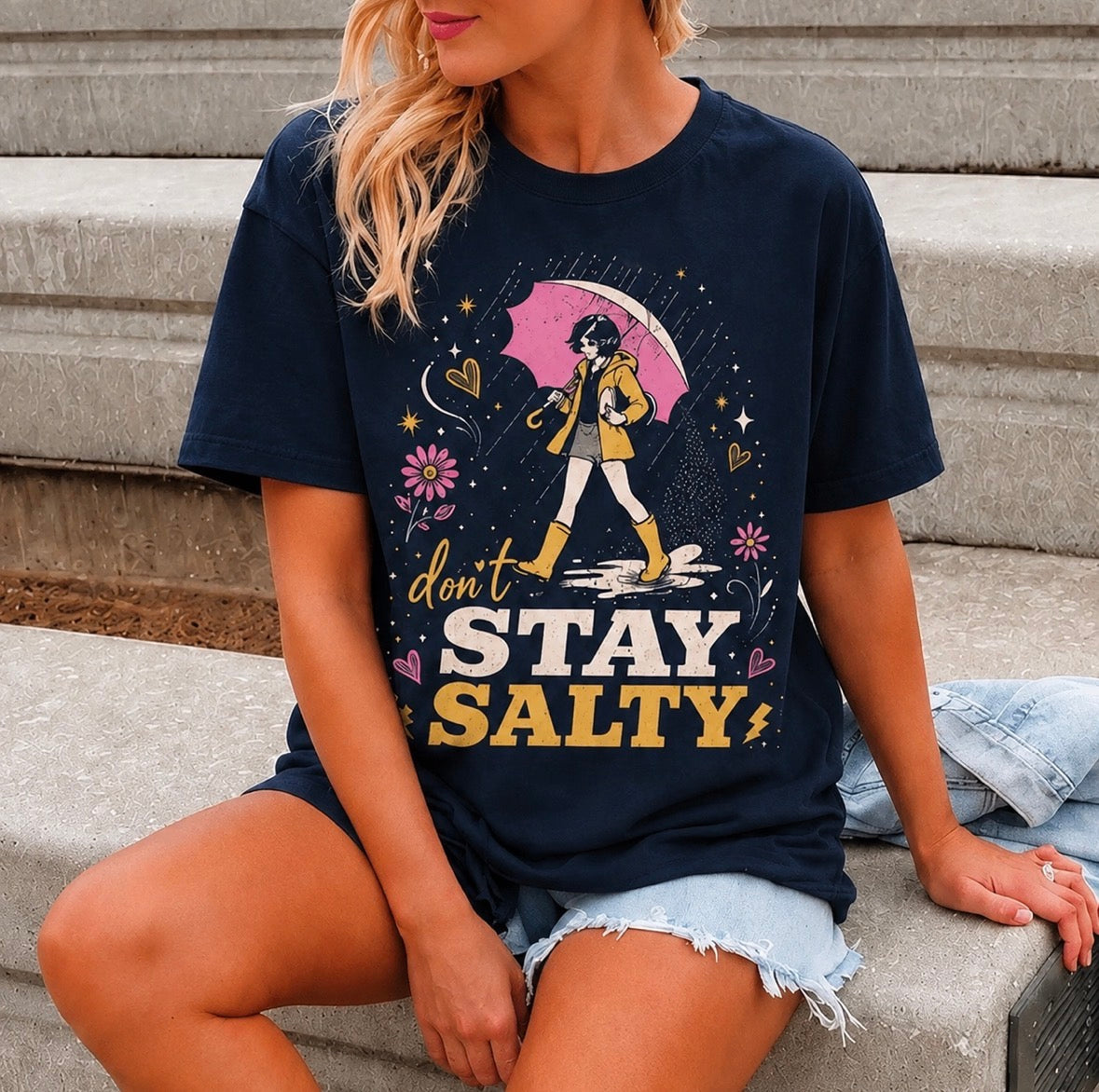 A woman sitting on concrete steps wearing a navy blue graphic t-shirt with a yellow, pink, and white 'don't STAY SALTY' design, paired with light denim shorts.