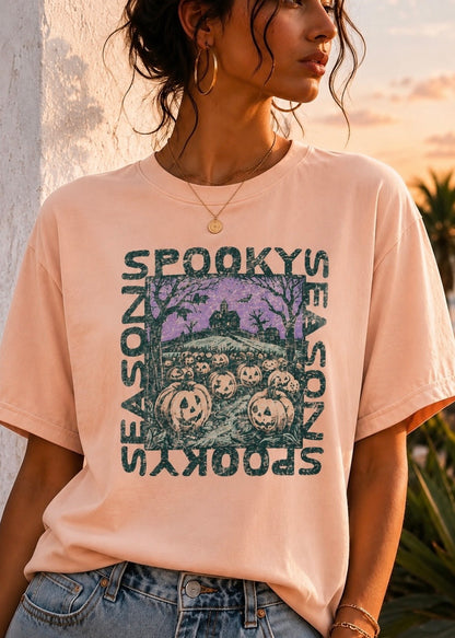 A woman wearing a light peach-pink graphic t-shirt with a distressed Halloween print that reads 'SEASON BY SEASON, SPOKY BY SPOKY' around a haunted house and jack-o'-lanterns. The photo is taken outdoors at sunset.