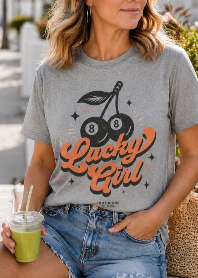 Woman wearing a gray t-shirt with 'Lucky Girl' design, holding a drink. Graphic includes cherry eight balls.