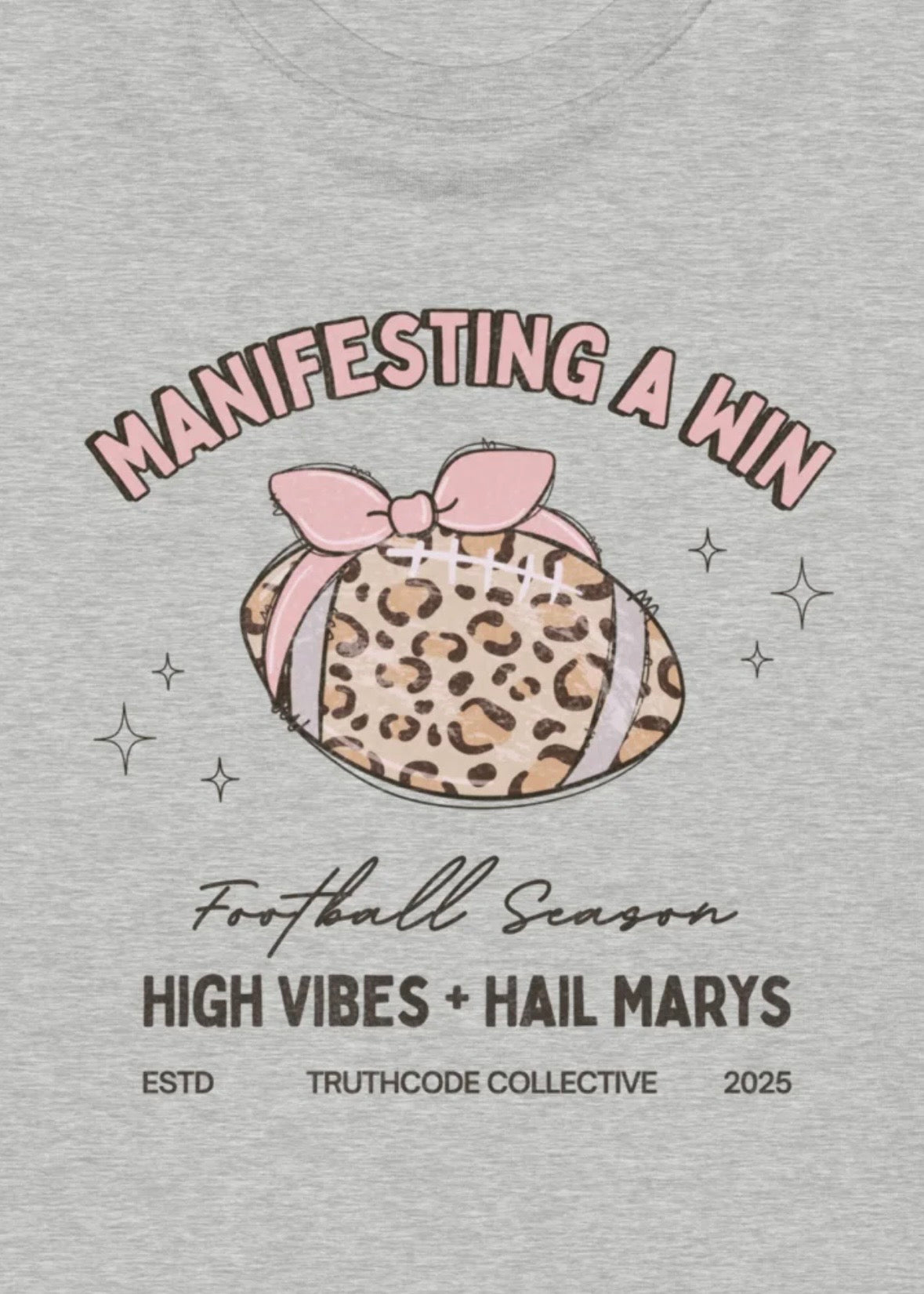 Close view of gray t-shirt with a leopard print football and pink bow graphic, with  text 'Manifesting a Win', 'Football Season', 'High Vibes + Hail Marys'