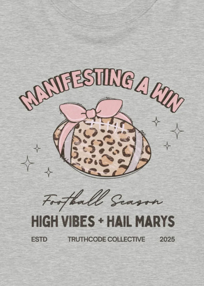 Close view of gray t-shirt with a leopard print football and pink bow graphic, with  text 'Manifesting a Win', 'Football Season', 'High Vibes + Hail Marys'