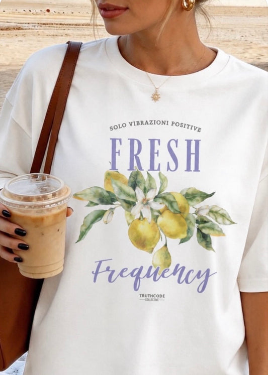 Person wearing a white t-shirt with lemon design and text 'fresh frequency', holding a drink.