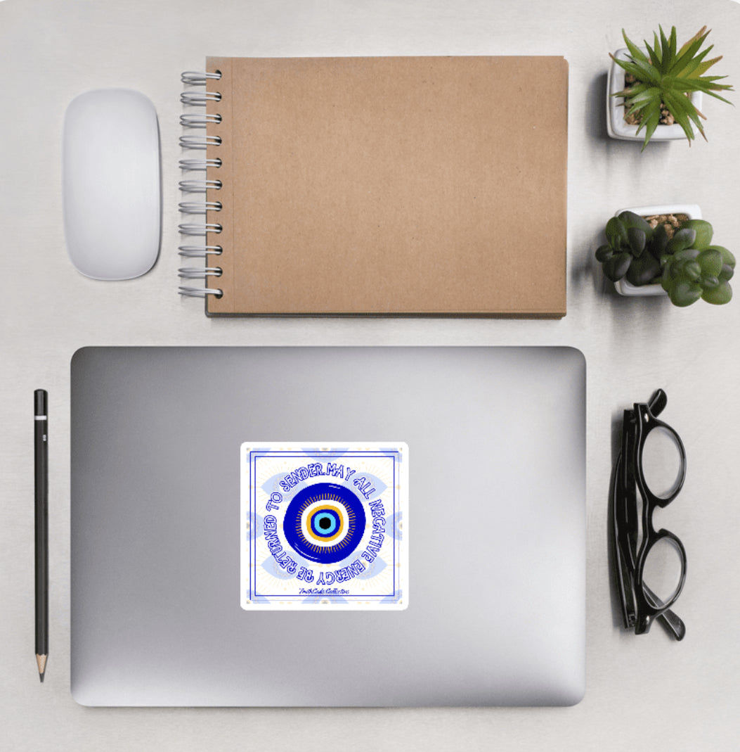 Aerial view of a desktop with laptop showing a squared sticker with eye design and text 'May all negative energy be returned to sender' on a white background
