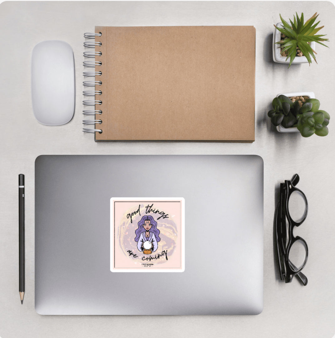 Aerial view of a desktop with laptop showing a squared sticker of a woman holding a magic ball and text 'Good things are coming' 