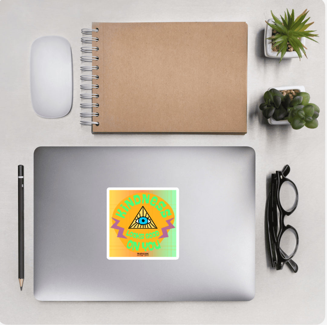 Aerial view of a desktop with laptop showing a colorful squared sticker with text 'Kindness looks good on you' and an eye and triangle design 