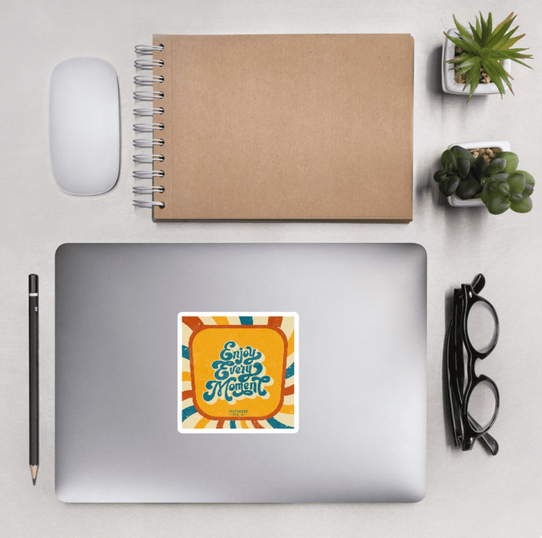 Aerial view of a desktop with laptop showing a squared sticker with a colorful vintage design and text 'Enjoy every moment'