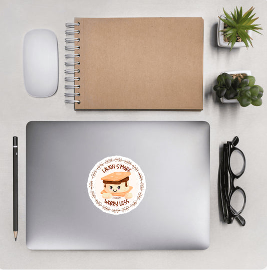 Aerial view of a desktop with laptop showing a circular sticker with a happy s'more design and text 'Laugh s'more worry less' 