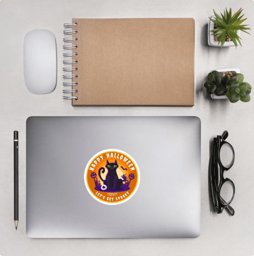 Aerial view of a desktop with laptop showing a circular sticker with black cat design and text 'Happy Halloween, let's get spooky'