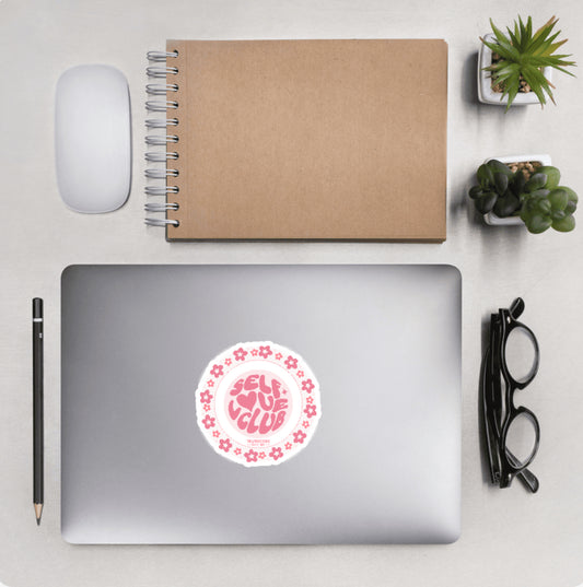 Aerial view of a desktop with laptop showing a circular sticker with pink flower border and text 'Self Love Club'