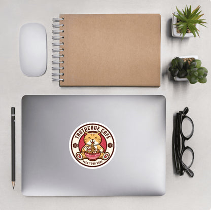 Aerial view of a desktop with laptop showing a circular sticker with a design of a cartoon cat eating noodles and text 'TruthCode Cafe, feed your soul'