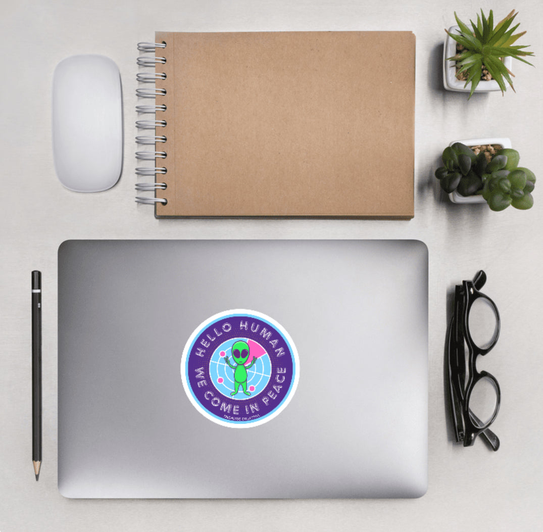 Aerial view of a desktop with laptop showing a circular sticker with a cute cartoon alien and text 'Hello human, we come in peace'