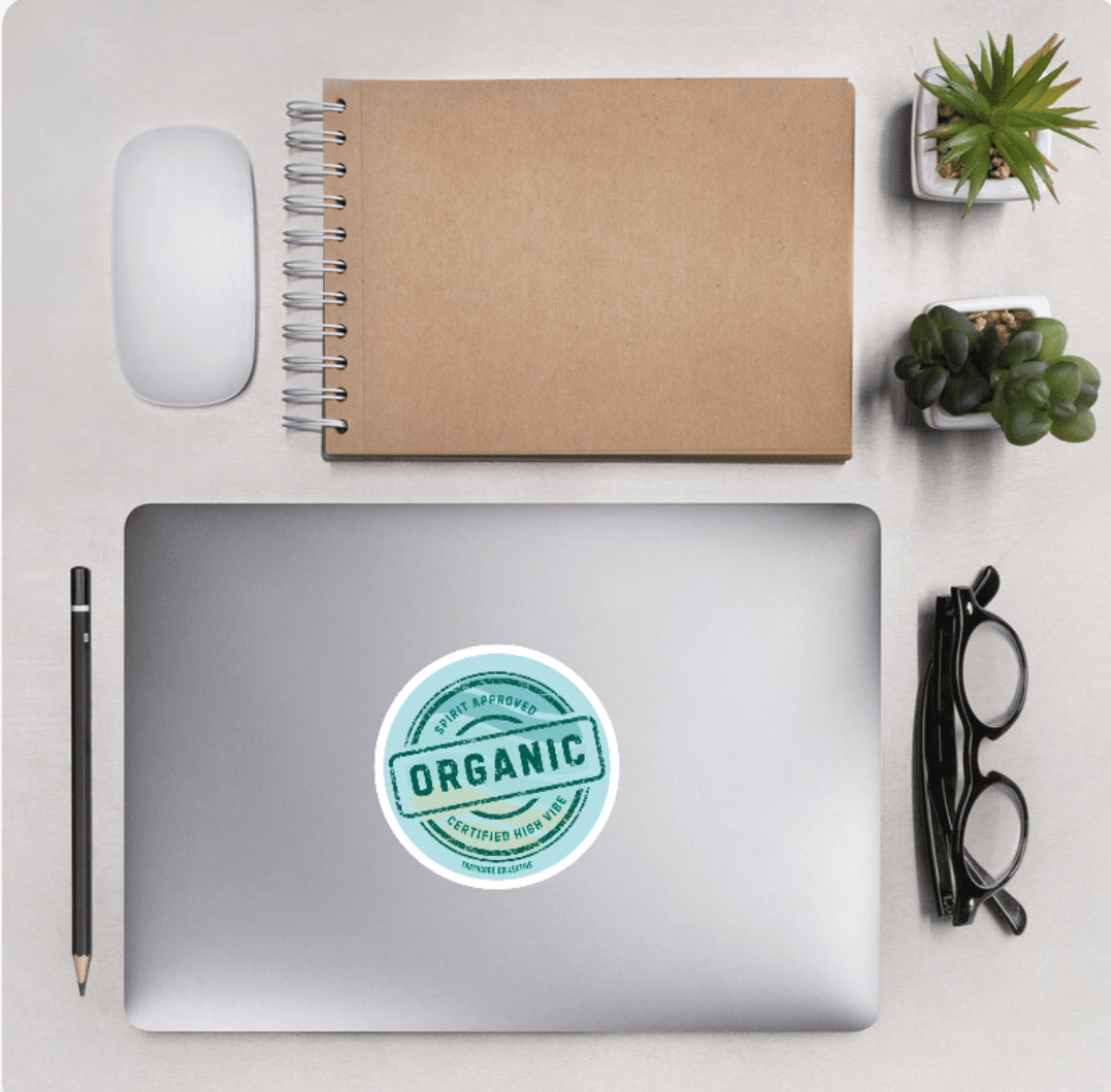 View showing desktop accessories and laptop with round sticker with text 'Organic' and Spirit approved, certified high vibe' on a light blue background