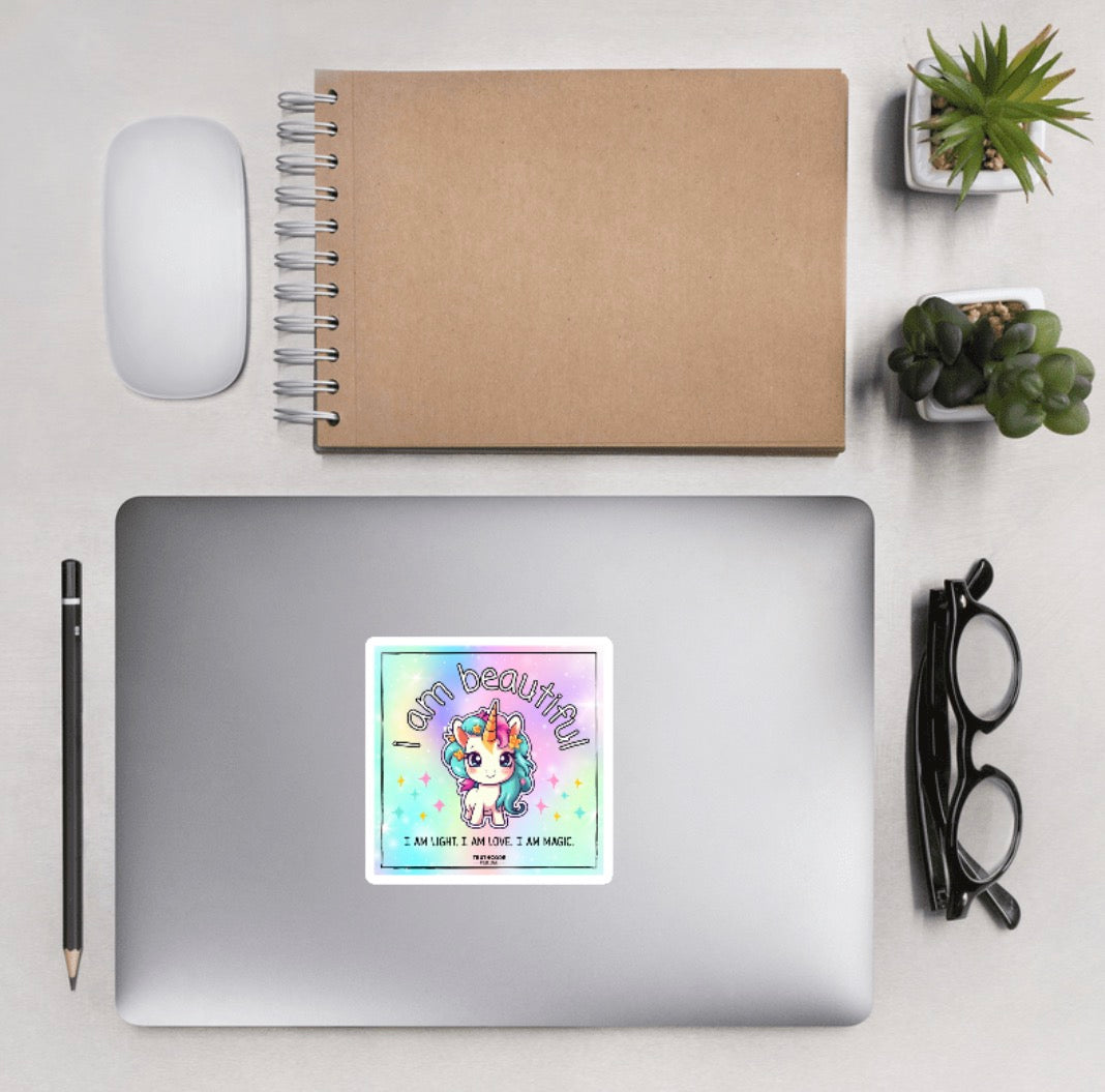 Aerial view of laptop with a sticker design of a unicorn and motivational text 'I am beautiful. I am light. I am love. I am magic.' on a colorful gradient background. Laptop is on a desk with accessories.
