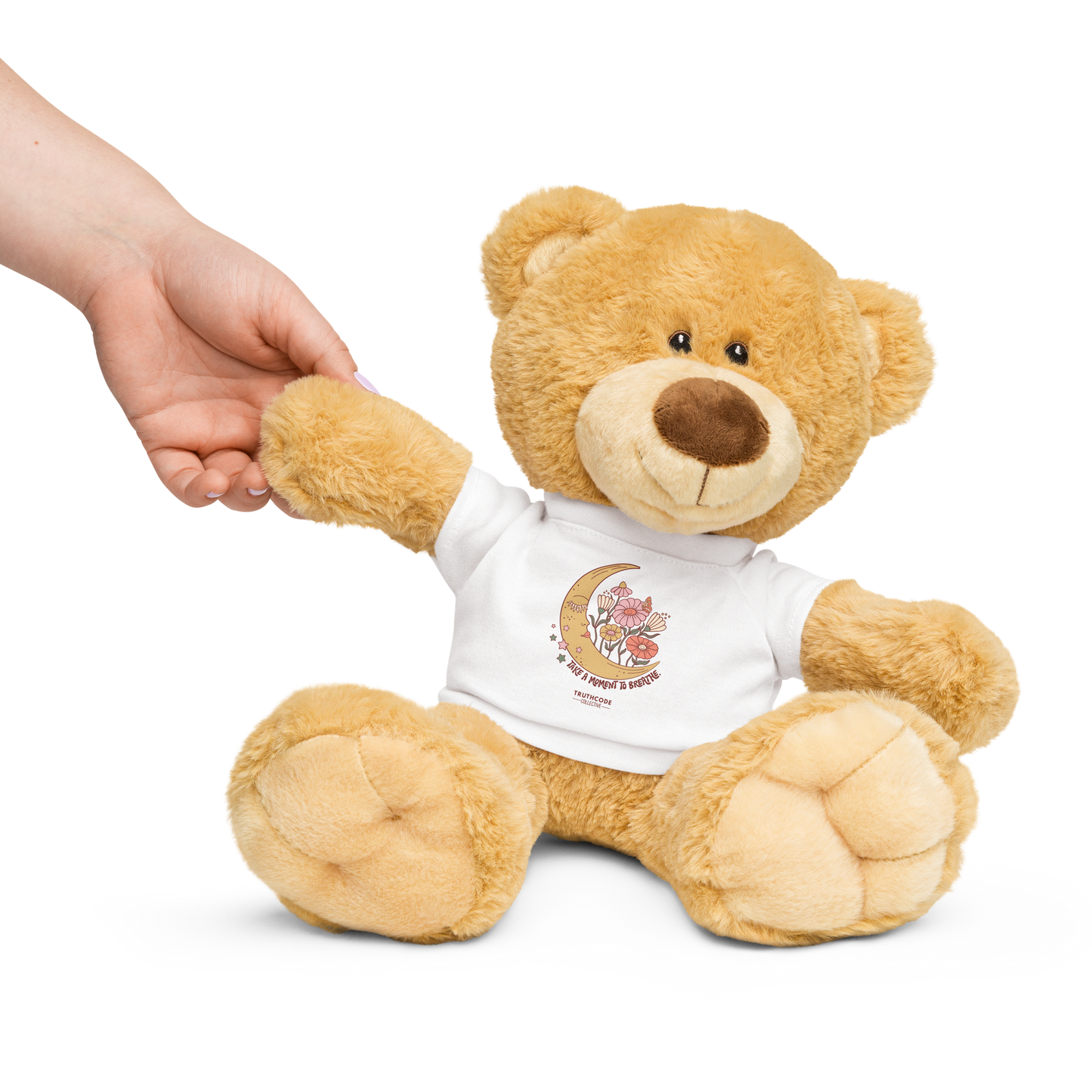 Teddy bear wearing a white shirt with a moon and flower design and text 'Take a moment to breathe', held by a hand on a white background