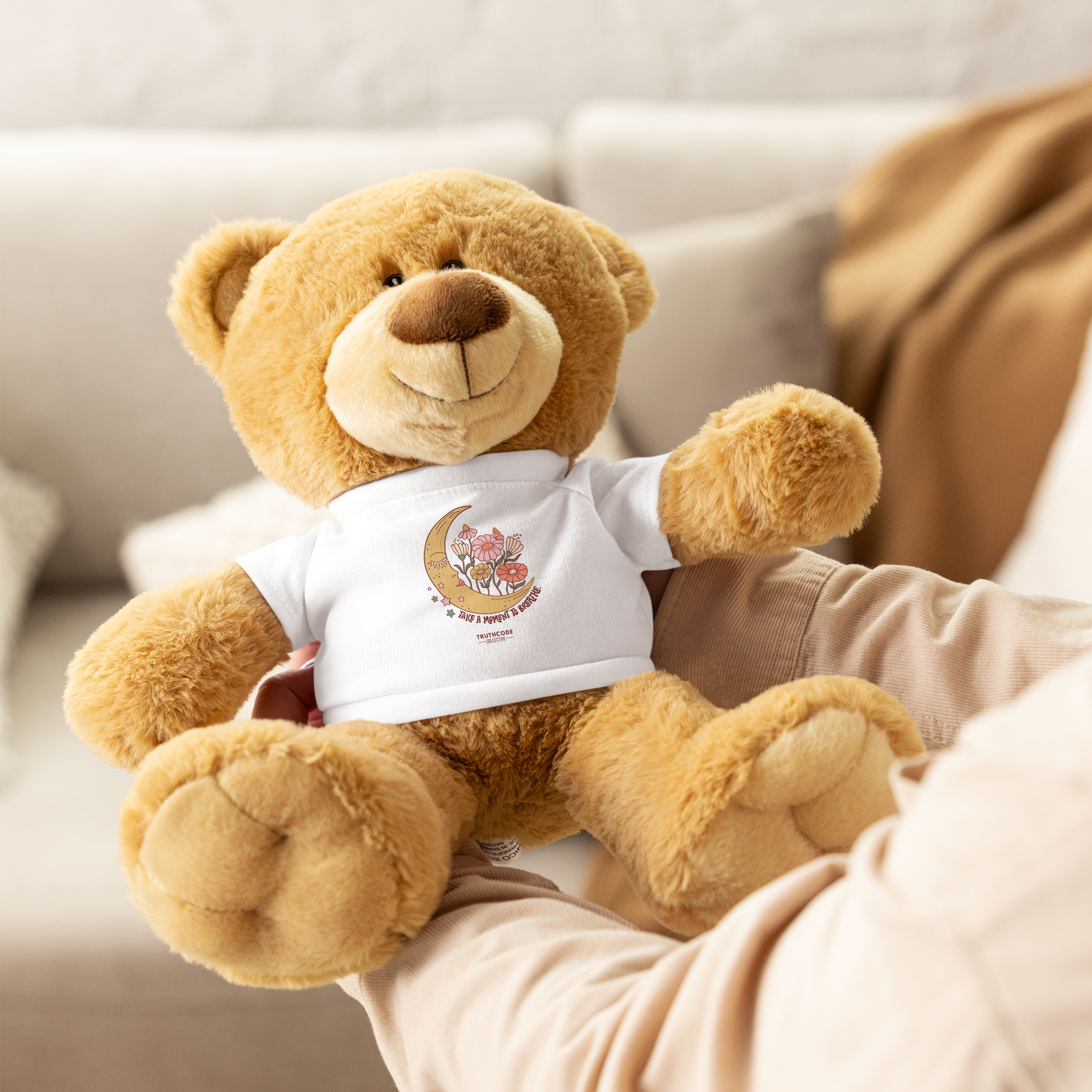 Tan teddy bear wearing a white shirt with a moon and flower design and text 'Take a moment to breathe', held by a person with a blurred background