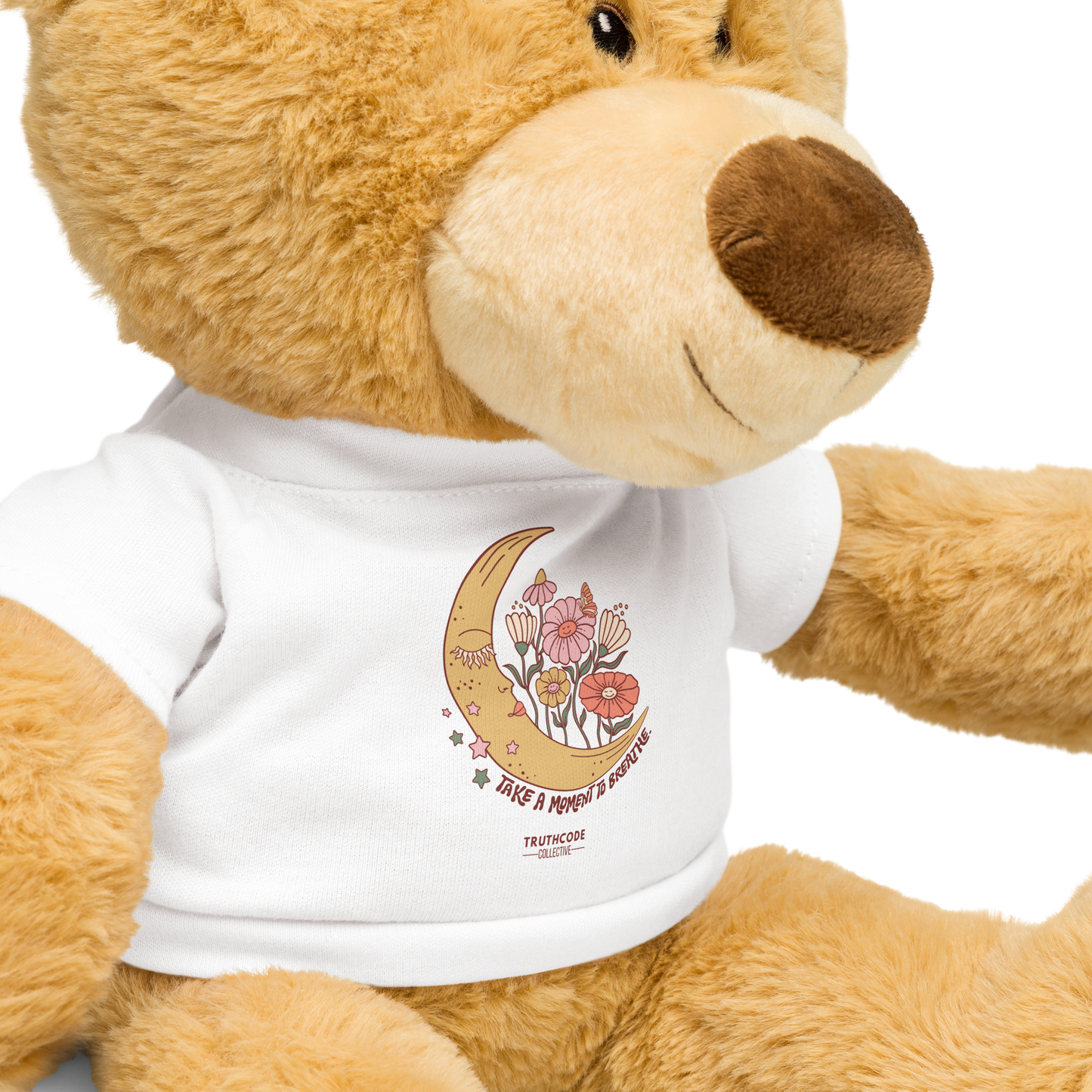 Close view of teddy bear wearing a white shirt with a moon and flower design and text 'Take a moment to breathe', on a white background