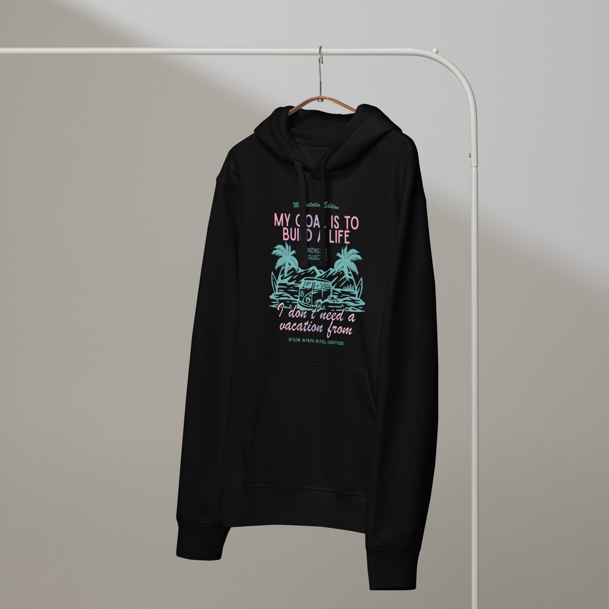 Black organic hoodie with tropical graphic and text 'My goal is to build a life I don't need a vacation from' on a hanger with a gray wall background