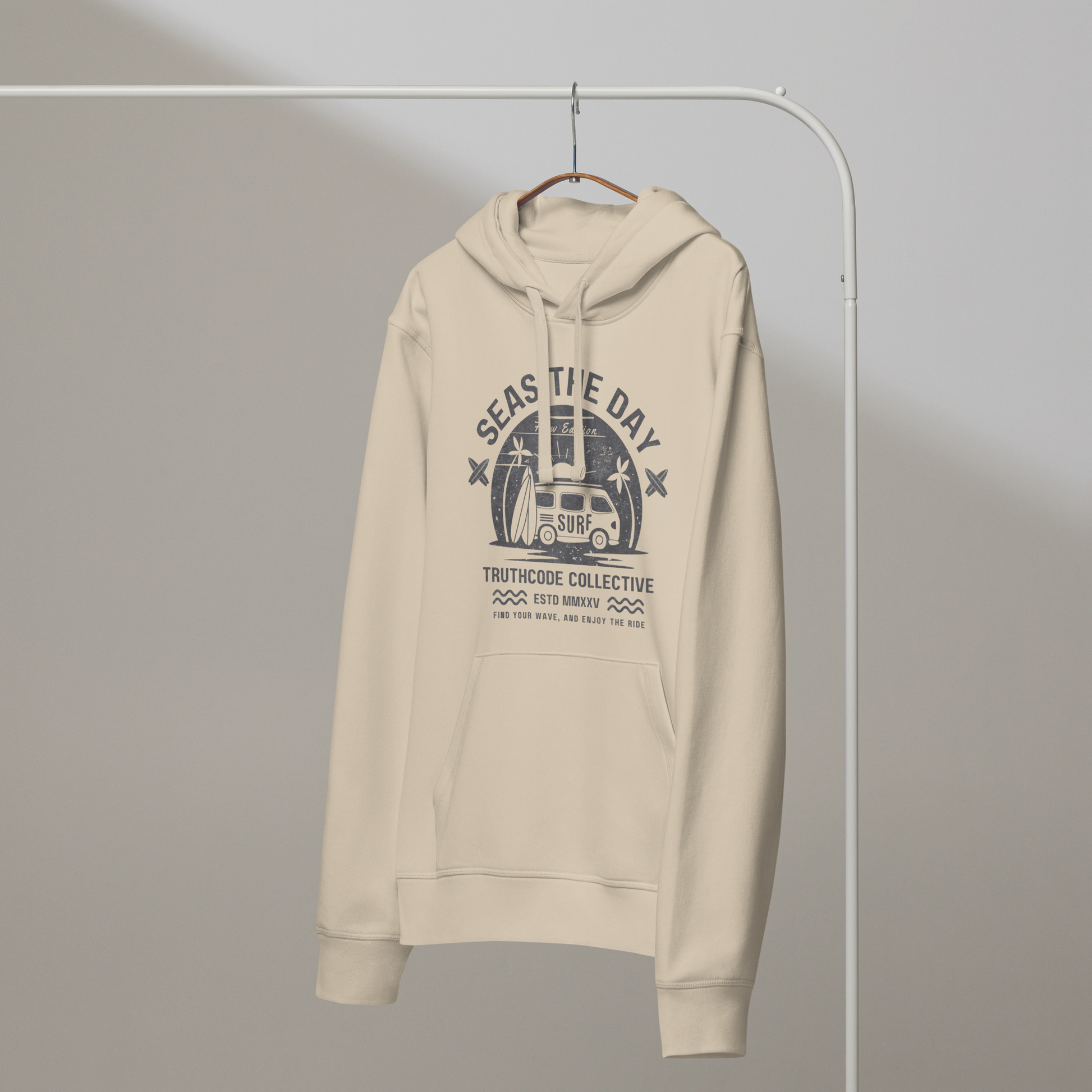Beige organic hoodie with graphic design of a vintage surfing van and text 'SEAS THE DAY' on a white hanger with a gray wall background