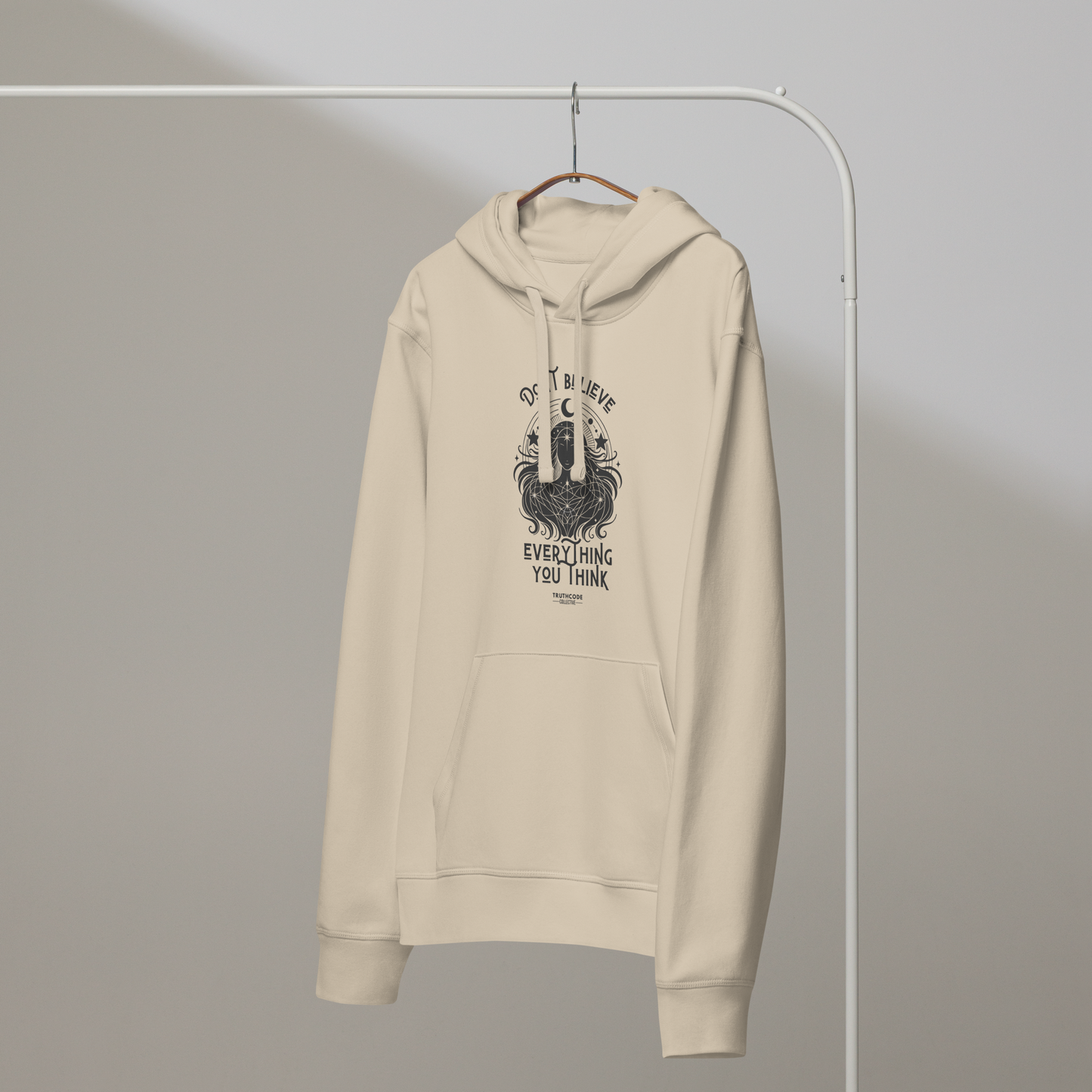 Beige organic hoodie with cosmic woman graphic design and text 'Don't believe everything you think' on a hanger with a gray wall background