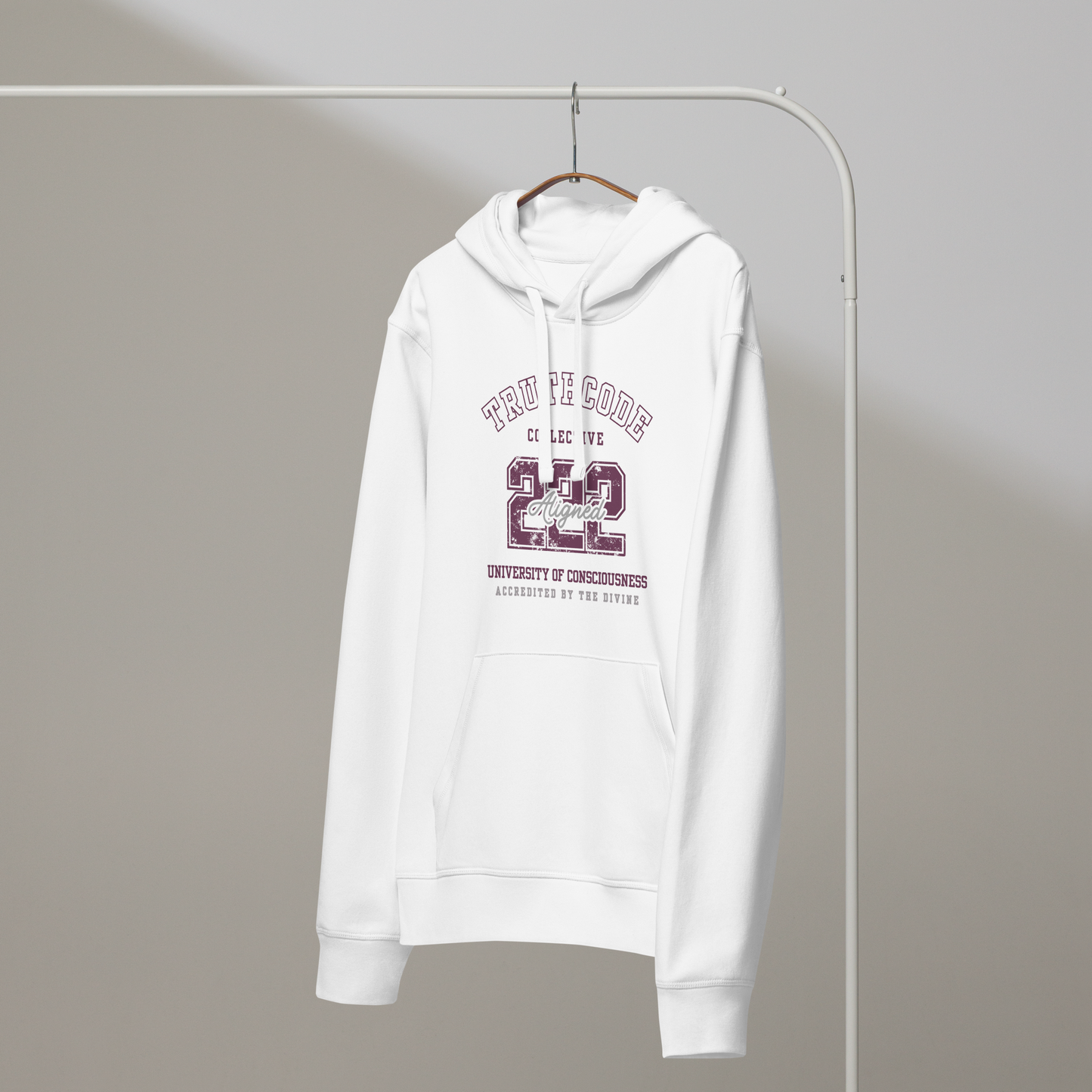 White organic hoodie with maroon text '222, Aligned' and 'University of consciousness, accredited by the divine'  on a white hanger with a gray wall background