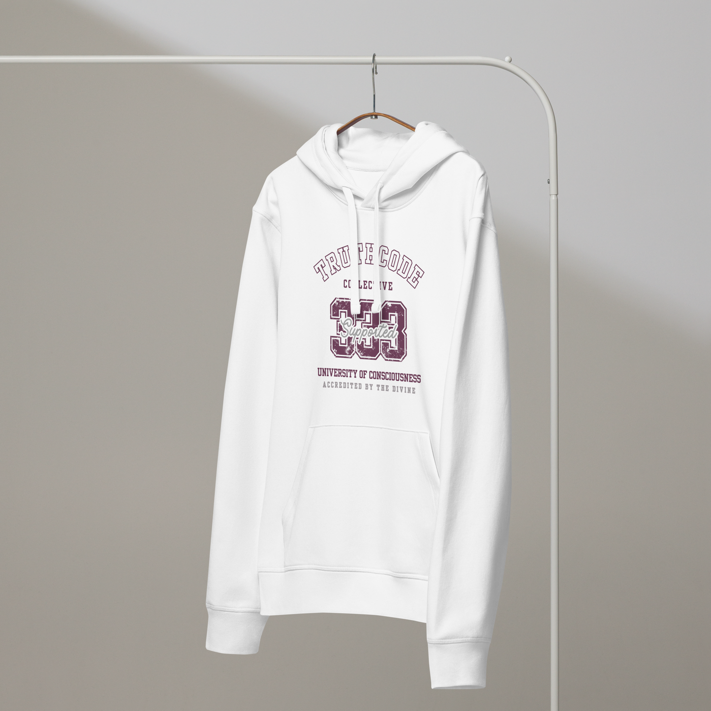 White organic hoodie with maroon text '333, Supported' and 'University of consciousness, accredited by the divine'  on a white hanger with a gray wall background