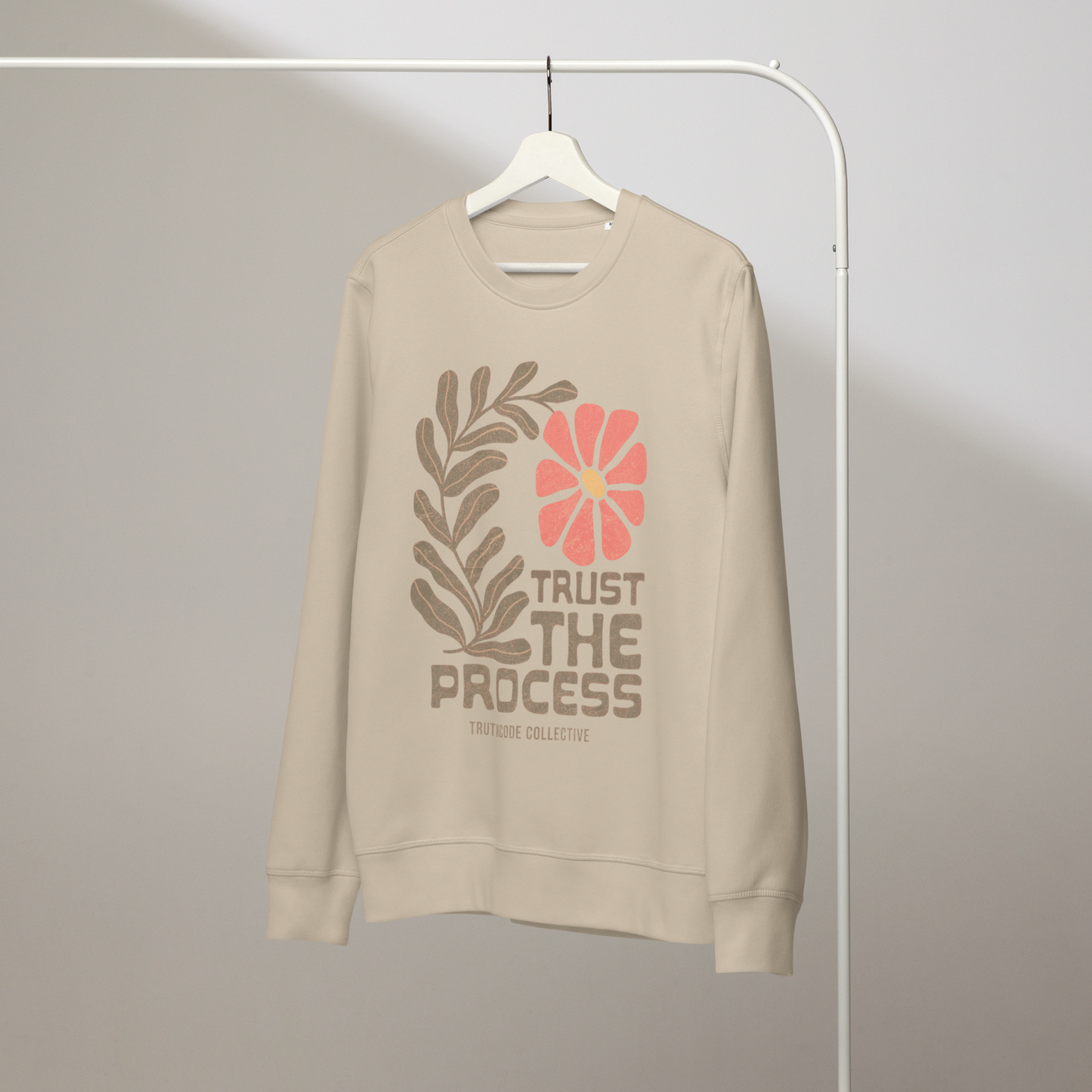 Beige organic sweatshirt with floral design and 'Trust The Process' text on a white  hanger with a gray wall background