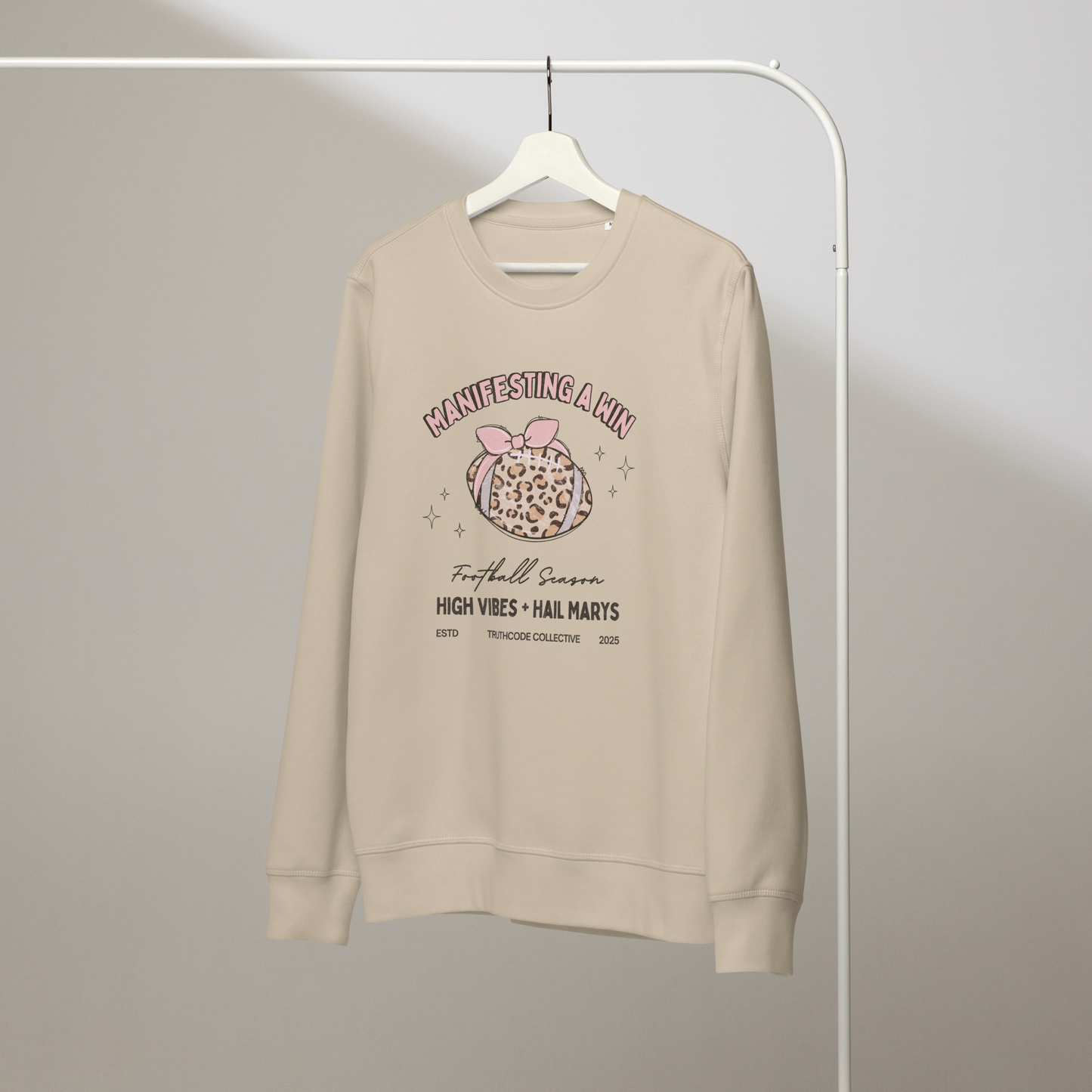 Beige organic sweatshirt with girly football graphic design and text 'Manifesting a win, high vibes + hail marys' on a white hanger with a gray wall background