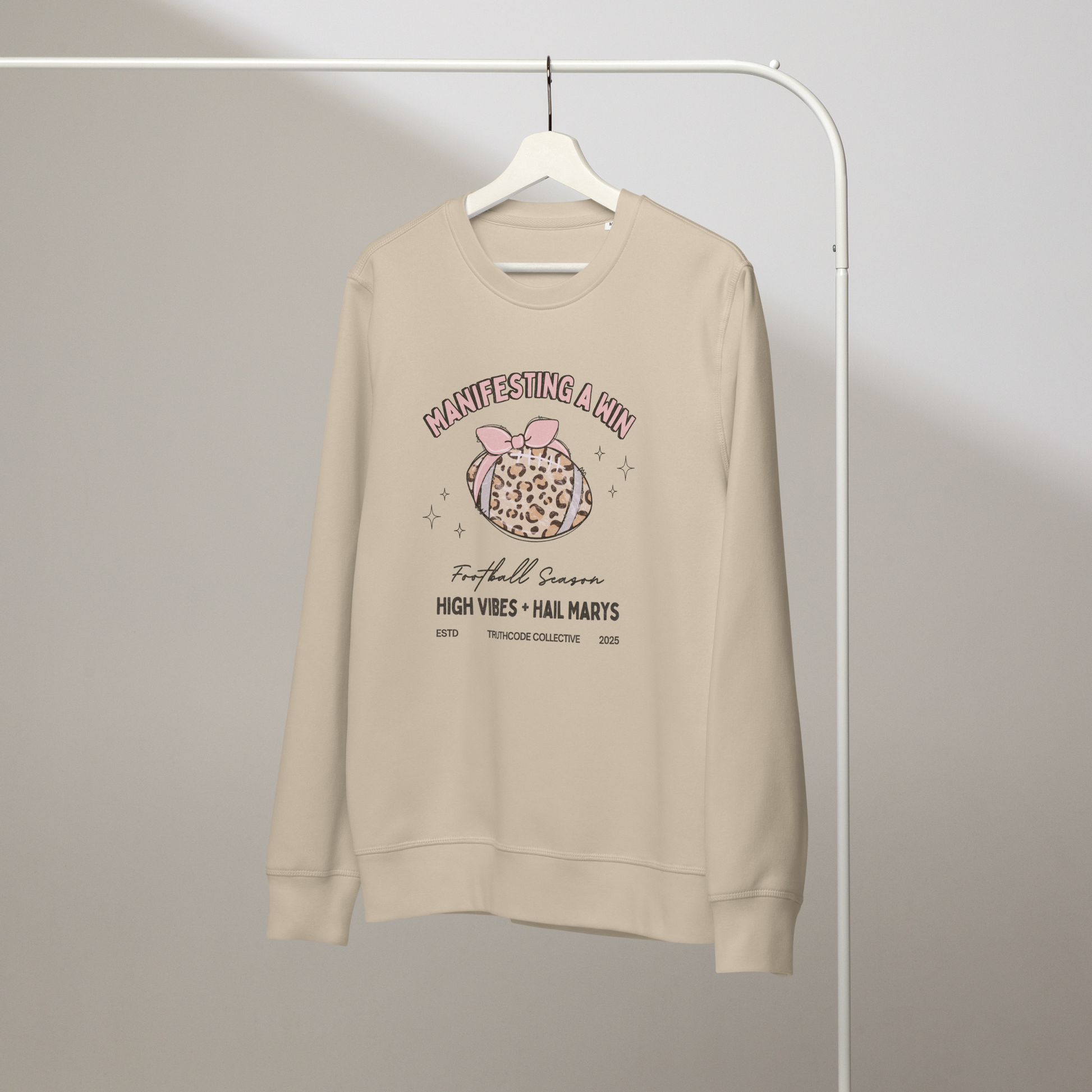 Beige organic sweatshirt with girly football graphic design and text 'Manifesting a win, high vibes + hail marys' on a white hanger with a gray wall background