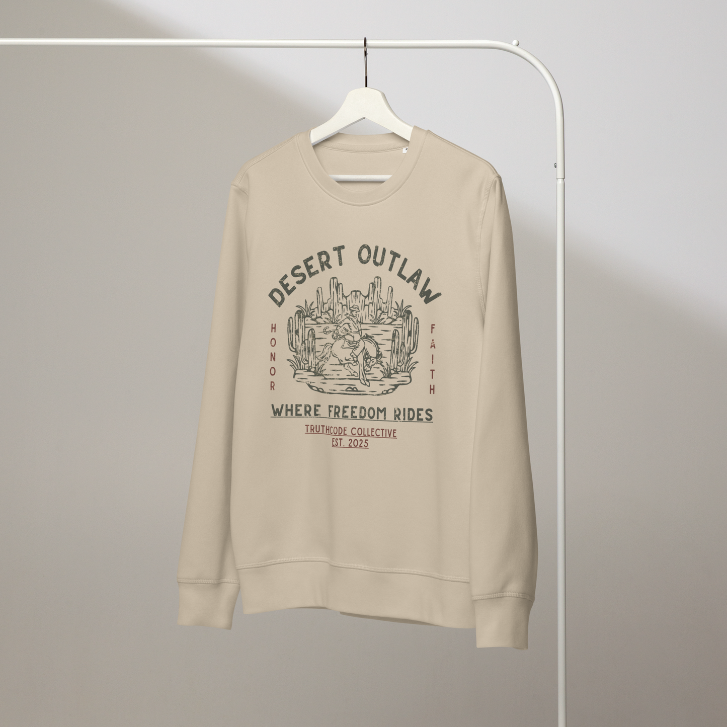 Beige organic sweatshirt with 'Desert Outlaw, where freedom rides' text and graphic design of cowboy riding a broco in the desert, on a white hanger with a gray wall background