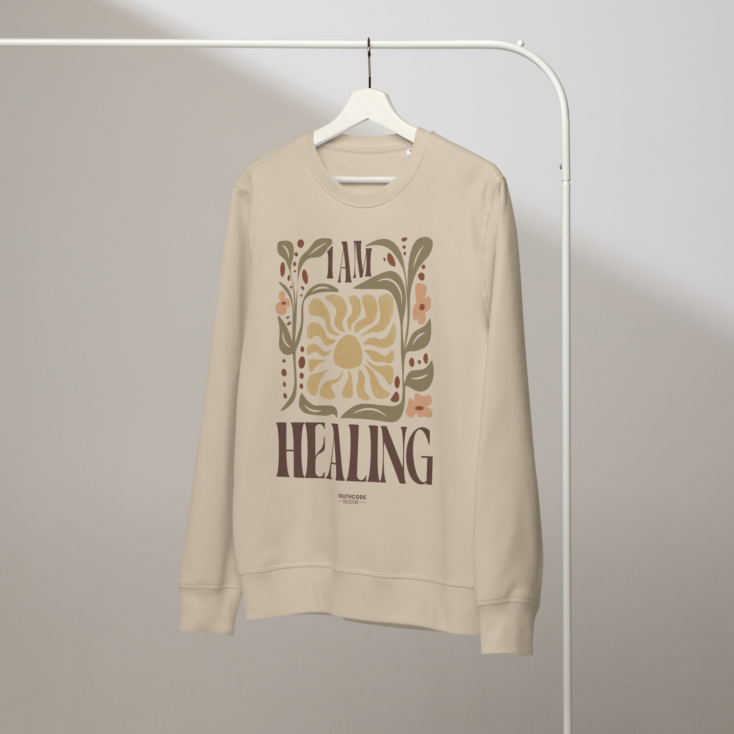 Beige organic sweatshirt with 'I AM HEALING' text and sun and plants design on a white hanger with a gray wall background