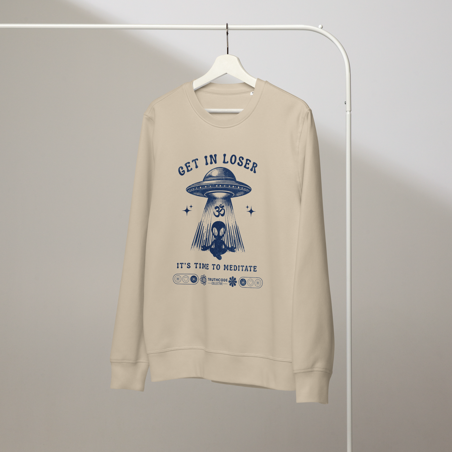 Beige organic sweatshirt with alien and spaceship graphic and text 'Get in loser, it's time to meditate' on a hanger with a gray wall background