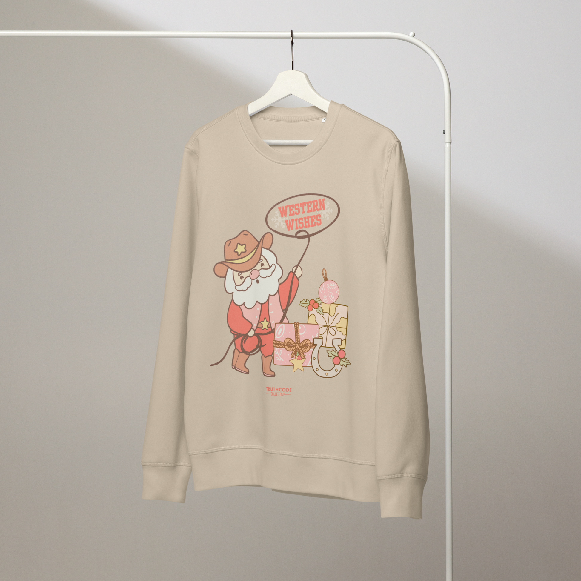 Beige organic sweatshirt with a cowboy Santa Claus graphic and 'Western Wishes' text on a hanger with a gray wall background