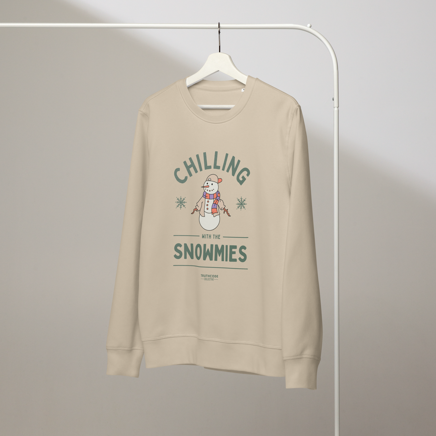 Beige organic sweatshirt with a snowman graphic and text 'Chilling with the Snowmies' on a hanger with a gray wall background