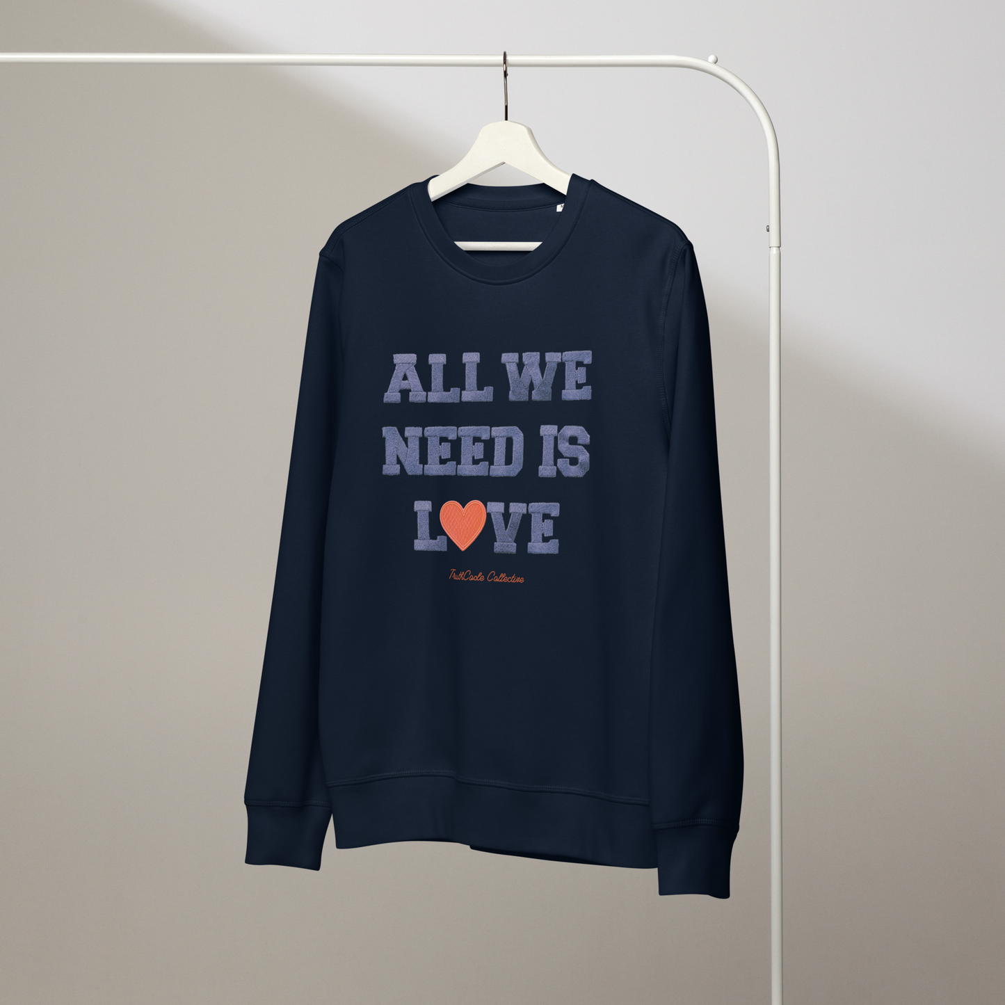 Navy blue organic sweatshirt with 'All We Need Is Love' text on a white hanger with a gray wall background