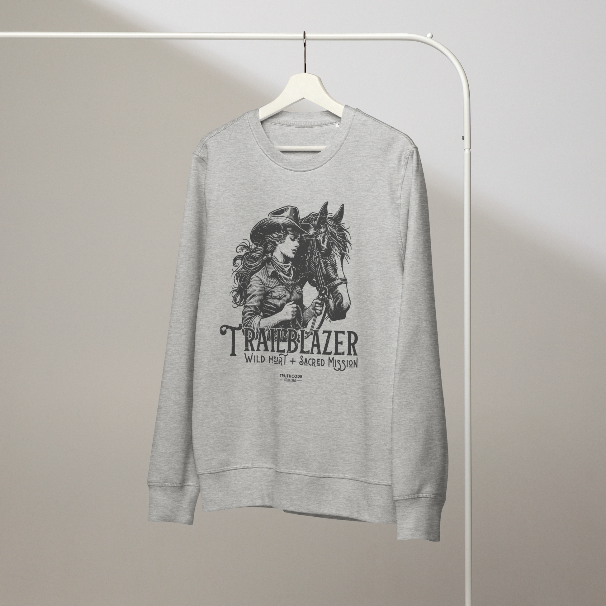 Gray organic sweatshirt with graphic of a woman holding a horse and text 'Trailblazer' and 'Wild heart, sacred mission' on a white hanger with a gray wall background